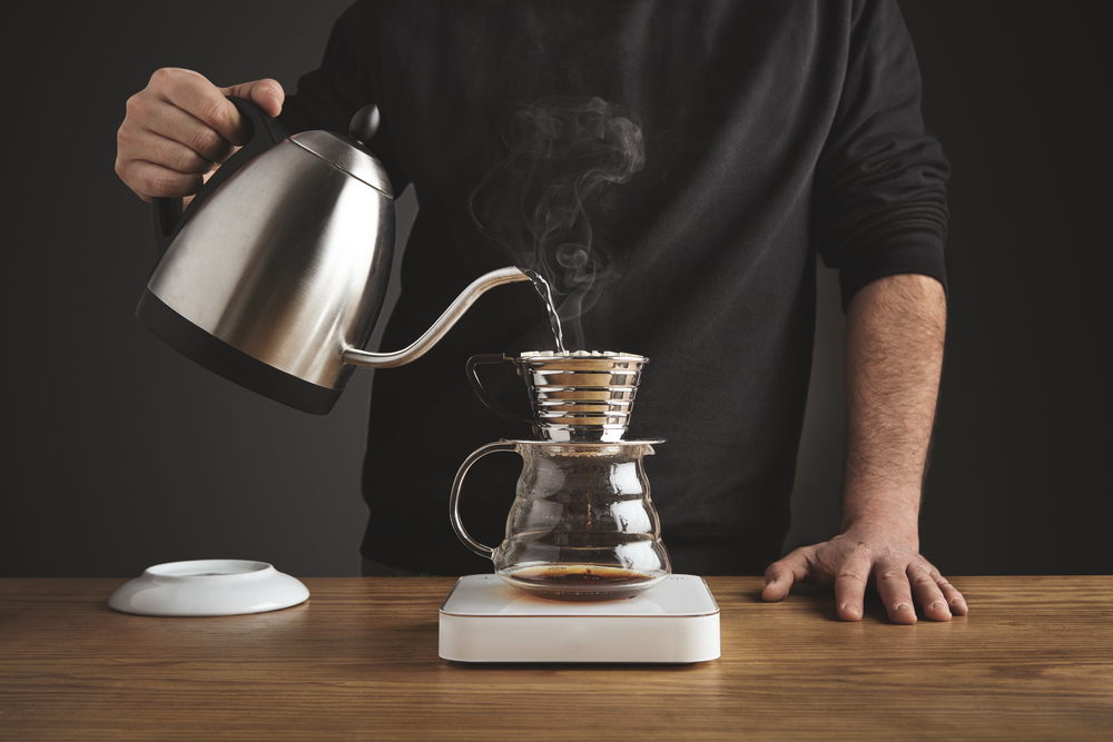 Barista in black sweatshot spills hot water to prepare filtered coffee from silver teapot to beautiful transparent chrome drip coffee maker on white simple weights. Everything on thick wooden table in cafe shop. Steam