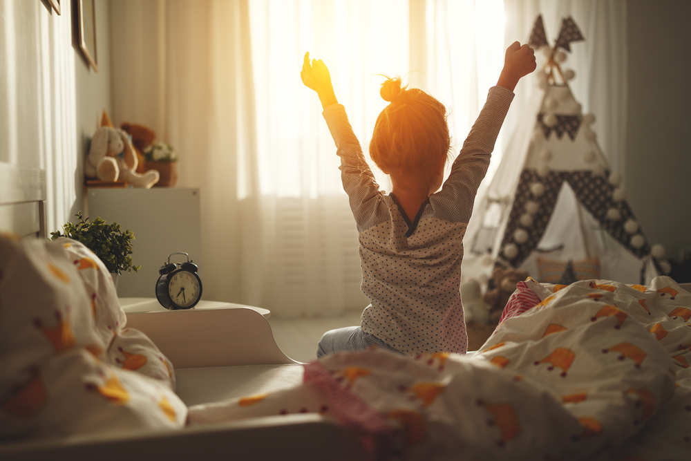 朝日が差し込む部屋のベッドの上で伸びをする小さな女の子