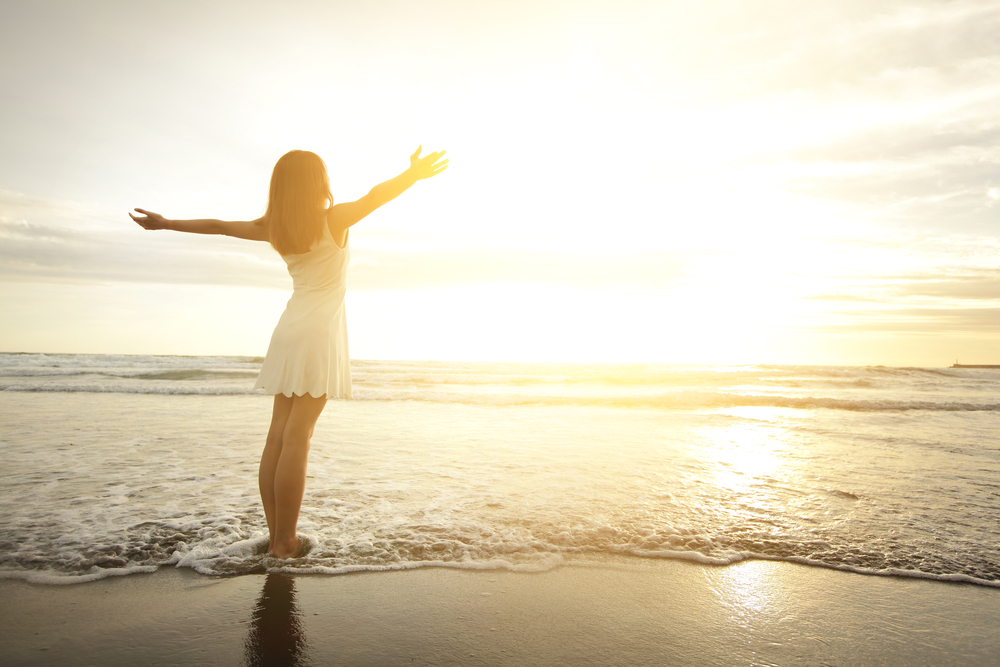 浜辺で両手を上げて沈む夕日を全身に浴びる若い女性